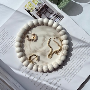 Puede incluir: Una bandeja decorativa redonda de color crema con un patrón similar al mármol, con joyas doradas: un par de pendientes de aro, un anillo y un collar de cadena. La bandeja tiene un borde con cuentas y está sobre una revista.