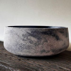 May include: A grey, textured bowl with a dark interior. The bowl has a rounded shape and sits on a wooden surface. The bowl's exterior has a mottled appearance, with variations in the grey tones. The overall aesthetic is rustic and minimalist.