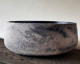 Puede incluir: Un cuenco gris texturizado con interior oscuro. El cuenco tiene forma redondeada y está sobre una superficie de madera. El exterior del cuenco tiene un aspecto moteado, con variaciones en los tonos grises. La estética general es rústica y minimalista.