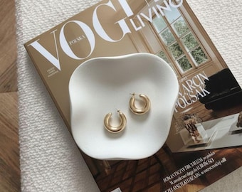 Peut inclure: Une paire de créoles dorées repose dans un plat blanc de forme organique, posé sur un magazine Vogue Living. La couverture du magazine est dorée avec le mot "POLSKA" visible. Les boucles d'oreilles sont de conception simple et classique.