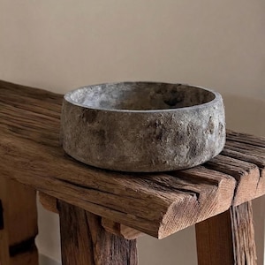 Puede incluir: Un cuenco redondo y poco profundo con una superficie gris texturizada y desgastada. El cuenco tiene un borde grueso y sólido y una cavidad interior profunda, mostrando un aspecto rugoso similar a la piedra con variaciones de tono.