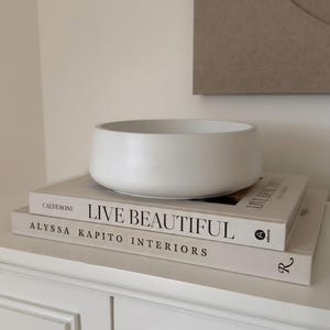 以下が含まれることがあります： 「Live Beautiful」と「Alyssa Kapito Interiors」というタイトルの2冊の本の上に、白い丸いセラミックボウルが置かれています。本は白いキャビネットに積み重ねられています。背景には、ニュートラルな色調の抽象的なアートワークが飾られています。