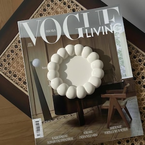 May include: A white, decorative, round tray with a bubble-like design sits atop a Vogue Living magazine. The magazine is open on a dark wooden table with a woven cane design. The tray is a modern home decor item.