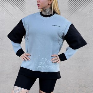 May include: A light blue sweatshirt with black sleeves and a white splatter pattern. The sweatshirt has a crew neck and the text "Don't Give Up" printed on the front.