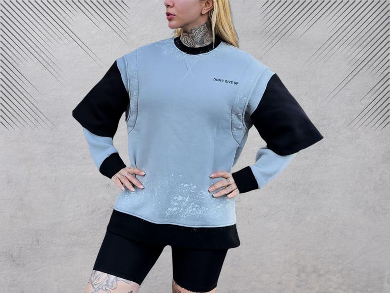 May include: A light blue sweatshirt with black sleeves and a white splatter pattern. The sweatshirt has a crew neck and the text "Don't Give Up" printed on the front.