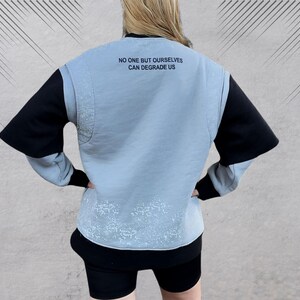 May include: A light blue sweatshirt with black sleeves and a white splatter pattern. The sweatshirt has the text "NO ONE BUT OURSELVES CAN DEGRADE US" printed on the back.