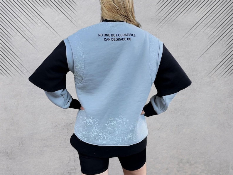 May include: A light blue sweatshirt with black sleeves and a white splatter pattern. The sweatshirt has the text "NO ONE BUT OURSELVES CAN DEGRADE US" printed on the back.