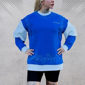 May include: A blue and light blue sweatshirt with a white graphic print. The sweatshirt has a crew neck and long sleeves.