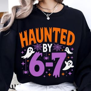 Puede incluir: Sudadera negra con las palabras "HAUNTED BY" en naranja, "6-7" en morado, y fantasmas, arañas y estrellas blancas. Diseño gráfico temático de Halloween.