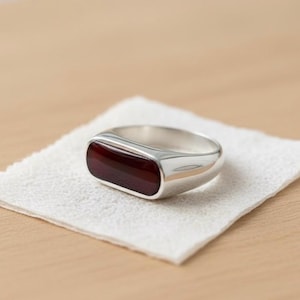 Puede incluir: Un anillo de plata con una piedra rectangular de color rojo oscuro. El anillo descansa sobre un paño blanco texturizado, sobre una superficie de madera marrón claro. El diseño del anillo es simple y moderno, con un acabado pulido.