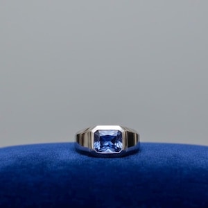 Puede incluir: Un anillo de plata con una piedra preciosa rectangular de color azul oscuro. El anillo está colocado sobre una superficie de terciopelo azul sobre un fondo gris. La piedra preciosa es el punto focal, mostrando un color vibrante y un diseño limpio y moderno.