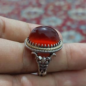 Pode incluir: Um anel de prata com uma grande pedra preciosa oval, translúcida, vermelho-alaranjada. A pedra preciosa é engastada em uma moldura detalhada com detalhes dourados e uma faixa de prata decorativa. O design do anel inclui enfeites ornamentados.