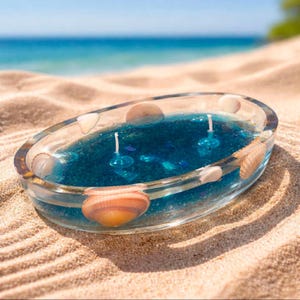 Peut inclure: Une bougie ovale en résine transparente avec un intérieur bleu profond et scintillant incrusté de divers coquillages. La bougie possède deux mèches blanches placées au centre de la cire bleue, créant un design inspiré du littoral.
