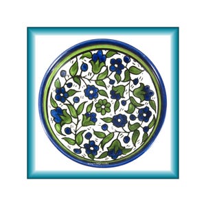 May include: A round ceramic plate featuring a white background with a hand-painted pattern of blue flowers, green leaves, and small blue berries. The plate has a thick blue rim and a thin green inner border. The text Belongs to Langer Jewelry is visible.