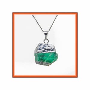 Op de afbeelding: Een ketting met hanger bestaande uit een ruwe, doorschijnende groene edelsteen met een textuurrijke, zilverkleurige metalen kap. De hanger hangt aan een dunne, zilverkleurige metalen ketting. De steen heeft een onregelmatige, grillige vorm met zichtbare interne insluitingen en een levendige smaragdgroene kleur.