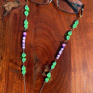 May include: A pair of black-framed spectacles attached to a decorative chain. The chain features green leaf-shaped beads, purple and white cylindrical clay beads, and gold-toned metal links. The spectacles have clear lenses and black temples.