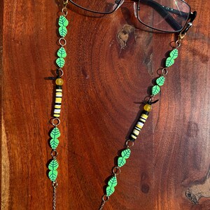 May include: A pair of black-framed spectacles attached to a decorative chain. The chain features green leaf-shaped beads, yellow translucent beads, and cylindrical beads with horizontal stripes in black, white, and yellow, connected by gold-tone metal links.