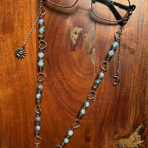 Könnte beinhalten: Brillenkette mit blauen und gelben Perlen, silbernen Herzakzenten und einem Sonnenanhänger. Die Kette ist an einer Brille mit schwarzem Rahmen befestigt. Der Text auf der Brille lautet: JERAULD 54018 142 C.