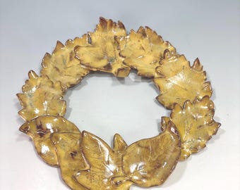 May include: A ceramic wreath with a leaf design. The leaves are a mix of yellow and brown, with a glossy finish. The wreath is circular and appears to be handmade.
