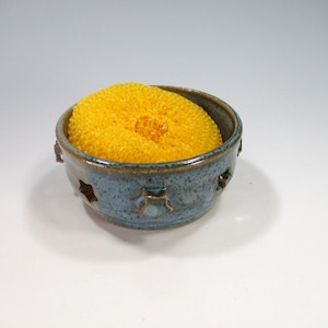 May include: A blue ceramic dish with cutouts, designed to hold a yellow sponge. The dish has a speckled glaze and a rustic look.