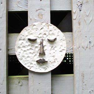 May include: A white, textured, circular face sculpture with closed eyes, a nose, and a mouth. The sculpture is mounted on a weathered, gray wooden fence. The face has a whimsical, handcrafted appearance.