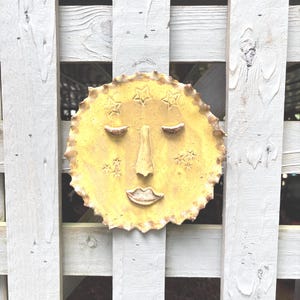 May include: A circular yellow ceramic wall plaque featuring a stylized sun face with closed eyes, a prominent nose, and full lips. The surface is speckled with brown dots and decorated with embossed star shapes around the facial features.