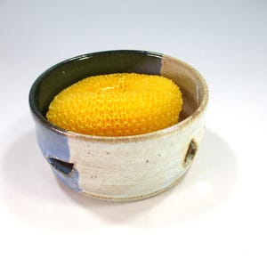 May include: A handmade ceramic bowl with a yellow sponge inside. The bowl has a white exterior with blue accents and a dark green rim. The sponge is a bright yellow color and has a textured, porous surface.