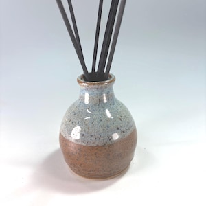 May include: A small, round ceramic diffuser vessel with a narrow neck. The bottom half features a speckled brown glaze, while the top half is finished in a speckled light blue glaze. Several thin, dark, straight reed sticks protrude from the opening.