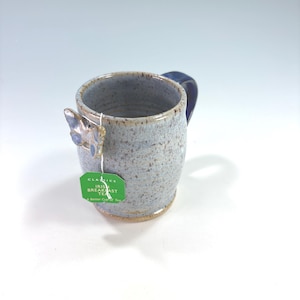 May include: A speckled light blue ceramic mug with a dark blue handle and a small butterfly-shaped accent on the rim. A green tea bag tag labeled Classics Irish Breakfast Tea A Better Cup of Tea hangs from the butterfly accent on the mug.