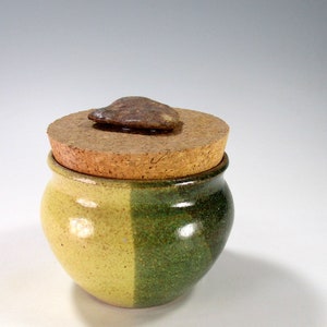 May include: A small, handcrafted ceramic jar with a cork lid. The jar is divided into two sections, one yellow and one green. A small, dark brown stone is affixed to the top of the cork lid. The jar is likely used for storage.