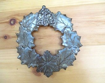 May include: A silver-colored Christmas wreath with holly leaves and a snowflake design. The wreath is made of metal and has a textured surface. A decorative bell is at the top of the wreath. The wreath is displayed on a wooden surface.