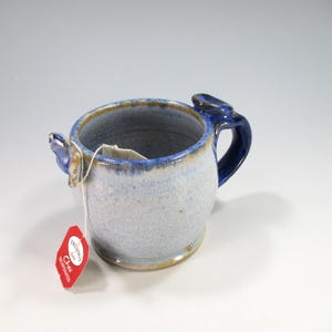 May include: A white ceramic mug with a black handle and the text "But First, Coffee" in a bold, black font. The mug is filled with a dark liquid, likely coffee, and sits on a light-colored surface.