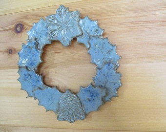 May include: A ceramic wreath with a light blue glaze. The wreath is shaped with stylized leaves and a snowflake design at the top. A bell shape is at the bottom. The wreath is displayed against a wooden background.