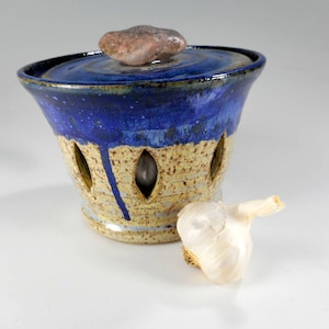 May include: A handmade ceramic garlic keeper with a blue and tan speckled design. The keeper has a lid with a natural stone handle and decorative cutouts. A whole garlic bulb rests beside the keeper.