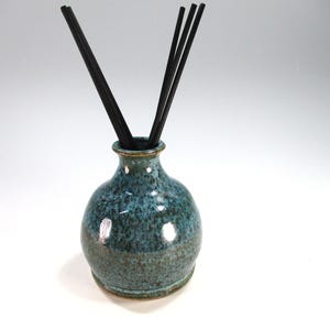 May include: A teal and brown ceramic essential oil diffuser with black reeds. The vase has a rounded body and a narrow neck. The surface has a speckled texture. The reeds are inserted into the vase.
