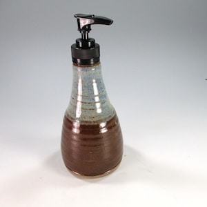 May include: A ceramic soap dispenser with a glossy finish, featuring a dark brown base that transitions into a light blue speckled neck. The vessel is topped with a black plastic pump mechanism designed for dispensing liquid soap or lotion.