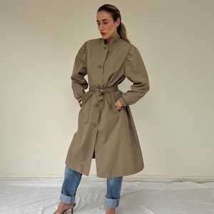 Op de afbeelding: Een beige trenchcoat met een hoge kraag, lange mouwen met geplooide boorden, een knoopsluiting aan de voorzijde en een bijpassende ceintuur in de taille. De jas wordt gecombineerd met een blauwe spijkerbroek en goudkleurige sandalen met hak.