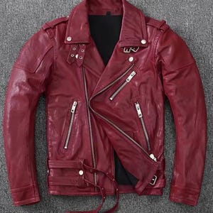 May include: A deep red leather jacket featuring a wide notched collar with silver-tone metal snaps, an asymmetrical front zipper closure, multiple zippered pockets, shoulder epaulets, and an adjustable buckled belt at the hemline.
