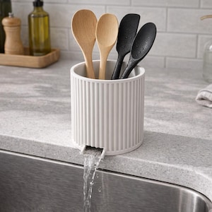 Op de afbeelding: Een witte, geribbelde keukengerei houder met een tuit, met houten lepels en zwarte spatels. Water stroomt uit de tuit in een roestvrijstalen spoelbak. Andere keukenartikelen zijn zichtbaar.