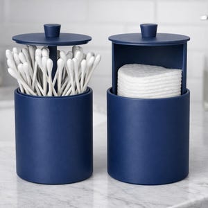 Può includere: Due contenitori da bagno blu navy con coperchi. Un contenitore contiene cotton fioc, l'altro dischetti di cotone. I contenitori cilindrici sono su un piano di lavoro in marmo. Un lavandino, un dosatore di sapone e asciugamani sono sullo sfondo.
