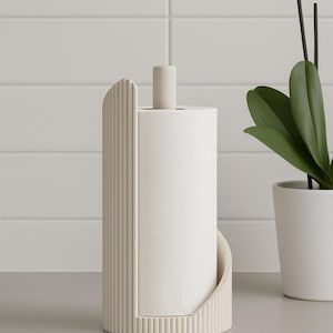 May include: A modern, off-white paper towel holder with a cylindrical design. The holder features a vertical, ribbed side panel and a small, cylindrical top piece. A roll of white paper towels is in the holder. A potted plant is in the background.