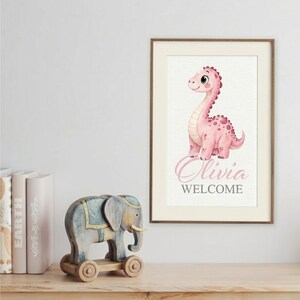 May include: A framed print featuring a pink dinosaur illustration with the name "Olivia" and the word "WELCOME" in cursive. A wooden elephant toy with wheels sits on a wooden surface.