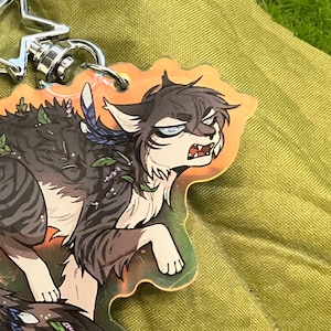 Puede incluir: Un llavero con un diseño de lobo de dibujos animados a rayas grises y negras, adornado con detalles florales. Tiene un cierre plateado en forma de estrella. La imagen está sobre musgo verde, una hoja y flores rojas.