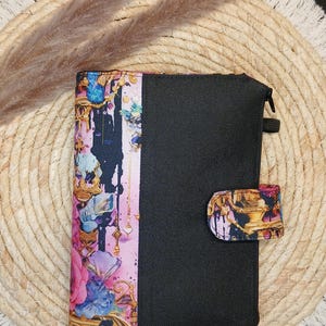 Könnte beinhalten: Eine rechteckige Stofftasche mit einem schwarzen Bereich auf der rechten Seite und einem lebendigen, mehrfarbigen abstrakten Blumenmuster mit Goldtönen auf der linken Seite. Die Tasche hat einen schwarzen Reißverschluss und eine passende Lasche.