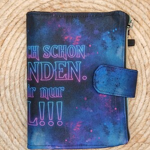 Könnte beinhalten: Ein rechteckiger Planer-Einband aus Stoff mit einem dunkelblauen und violetten Galaxie-Druck und verstreuten rosa Punkten. Die Vorderseite zeigt Text in einer Neon-Schriftart CH SCHON NDEN. ir nur L!!! sowie einen passenden Stoffverschluss.