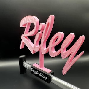 May include: A pink 3D-printed sign spelling "Riley" in a decorative font. The sign is mounted on a black base and supported by a silver rod with the text "Dazzle Clips". The background is black.