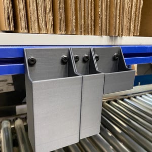 May include: Three gray storage bins attached to a blue and white shelf. The bins are rectangular and mounted with black screws. Cardboard boxes are stacked on the shelf above the bins. The bins are designed for organizing items in a warehouse or storage setting.