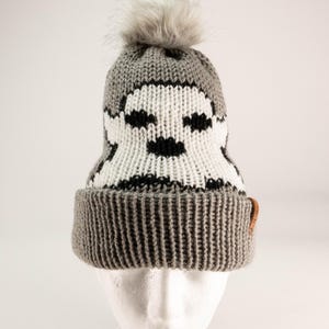 May include: A grey knit beanie with a white and black ghost graphic on the front. The hat features a ribbed cuff, a grey faux fur pom-pom on top, and a small brown rectangular tag attached to the side of the folded brim.