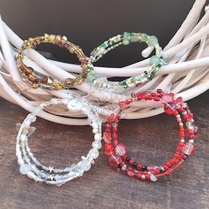 Pode incluir: Quatro pulseiras de contas enroladas em ouro, verde, branco e vermelho. As pulseiras são feitas com várias contas e cristais e são enroladas em torno de um fio central. As pulseiras são exibidas em uma superfície de madeira.