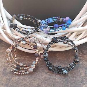 Pode incluir: Quatro pulseiras de contas em espiral em várias cores. Uma pulseira é preta, outra azul e as outras duas são castanhas e pretas. As pulseiras são feitas com pequenas contas e contas de destaque maiores. As pulseiras são exibidas em um fundo branco tecido.